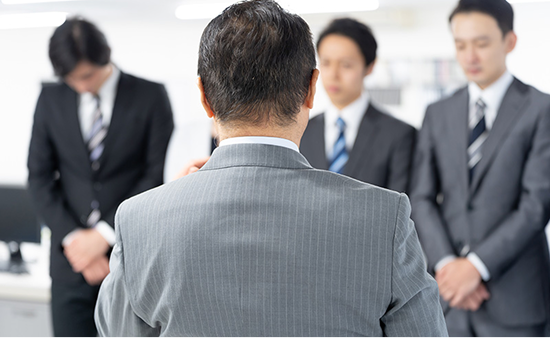 A senior businessman in a suit addresses three younger employees who are clasping their hands and looking down, reflecting workplace hierarchy.