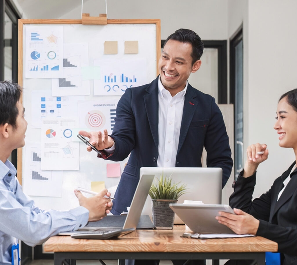 Business meeting scene with a man giving a presentation to two people, with charts and laptops on the table.