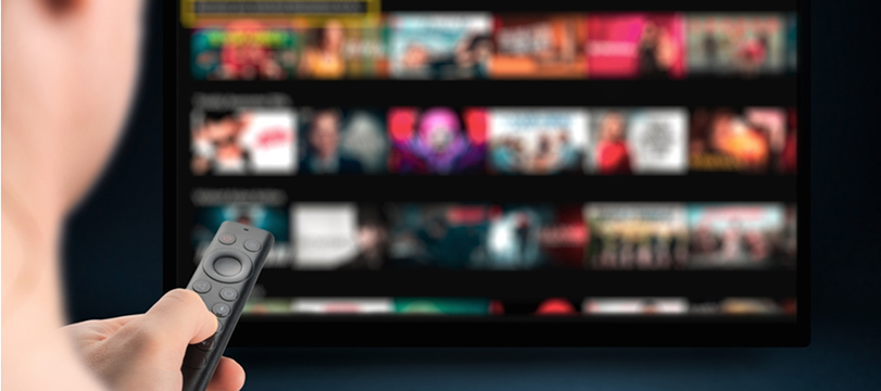リモコンを手にテレビで映像配信サービスを操作する人。画面には多くの映画や番組のサムネイルが並んでいる。